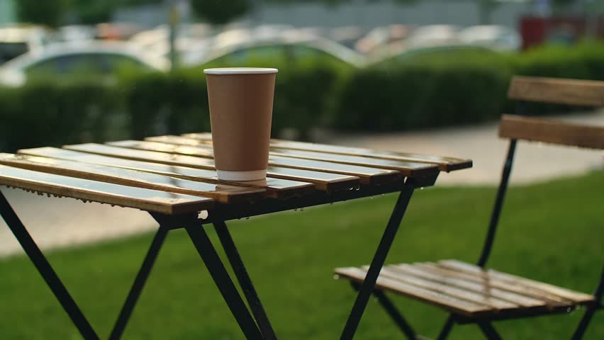 Rain drops dripping on paper coffee cup. Wooden tables of open terrace under rain. Street cafe in summer rain, slow motion