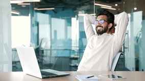 Young bearded businessman wearing glasses finished work on laptop while sitting at workplace in modern office. Happy handsome male stretches in a chair, throwing his hands behind his head. Work done - Powered by Shutterstock - Get 15% off with code: PIKWIZARD15