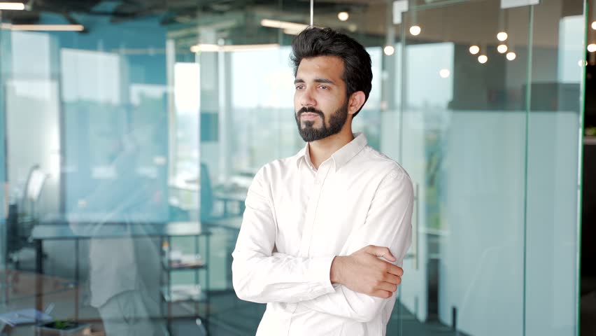 Portrait of a young successful bearded man in a shirt standing with crossed arms in a modern office. Confident smiling businessman with a friendly look looking at the camera. Head shot of an employee Royalty-Free Stock Footage #1107972349