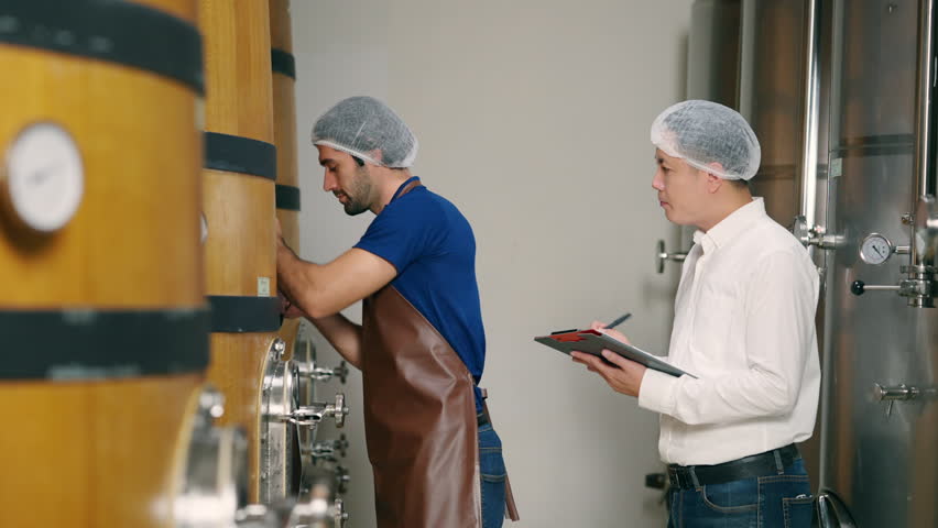 4K Professional male winemaker staff working and inspecting red wine quality in wine cellar with stainless steel barrels. Winery, wine shop, brewery liquor manufacturing industry business concept.