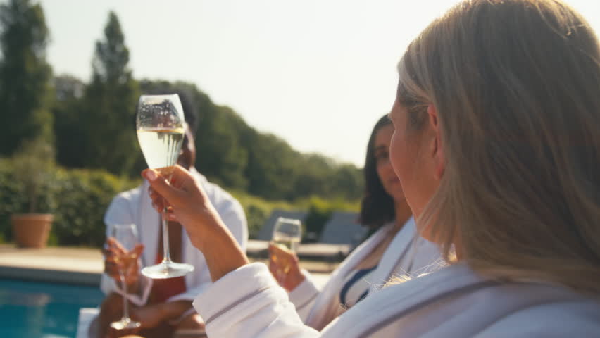 Three mature female friends wearing robes sitting outdoors on loungers by swimming pool drinking champagne and making a toast on spa day - shot in slow motion Royalty-Free Stock Footage #1107984701