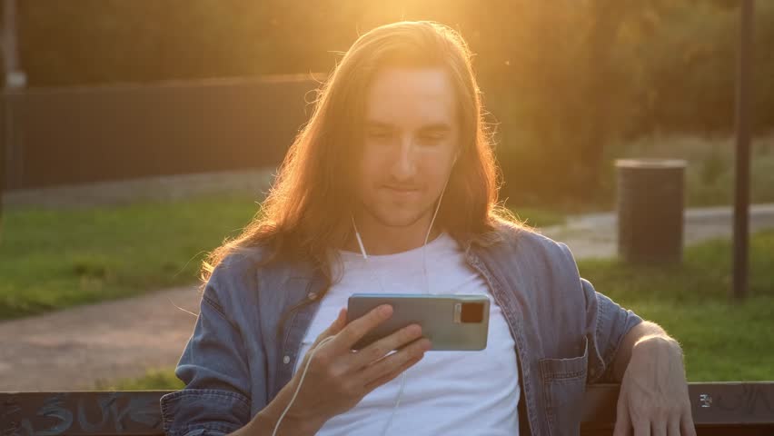 Young man watching video on the mobile phone while sitting at public park | Shutterstock HD Video #1107990965