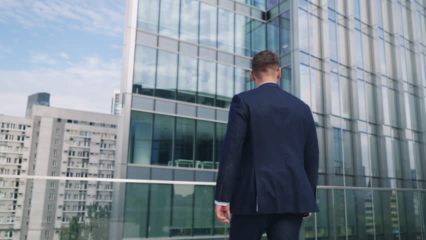 Relaxed male CEO businessman in suit standing on rooftop glass terrace in office building overlooking modern city downtown with high-rise skyscrapers on sunny day. Business people, gimbal slow motion Royalty-Free Stock Footage #1107995047
