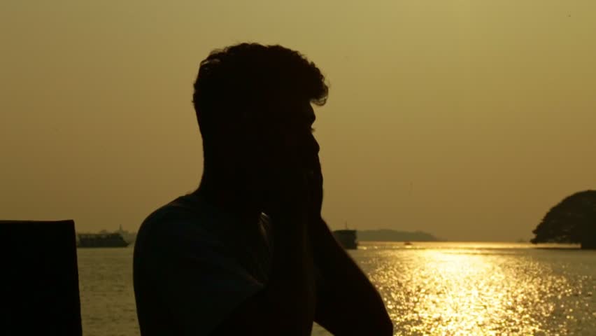 An Indian man leans against the wall beside the lake ,Sunset time , Houseboats in the background , Silhouette