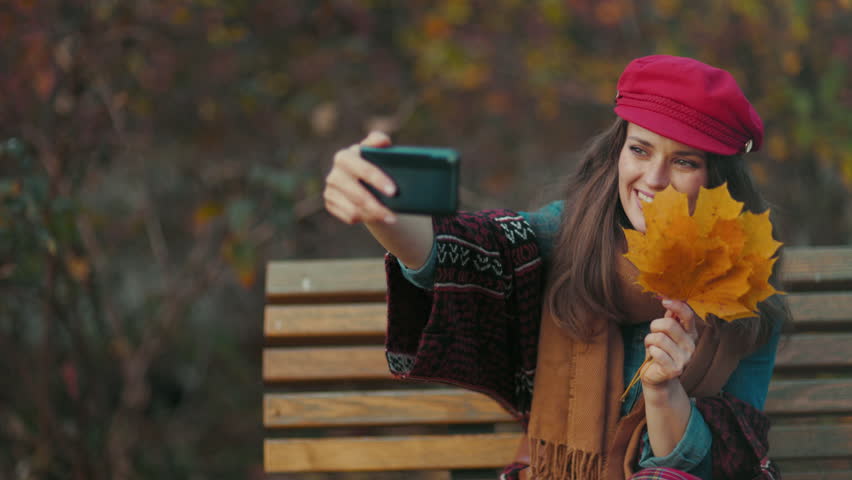 Hello autumn. smiling modern middle aged woman in red hat with autumn leafs and scarf having video meeting while sitting on bench in the city park. Royalty-Free Stock Footage #1108002207