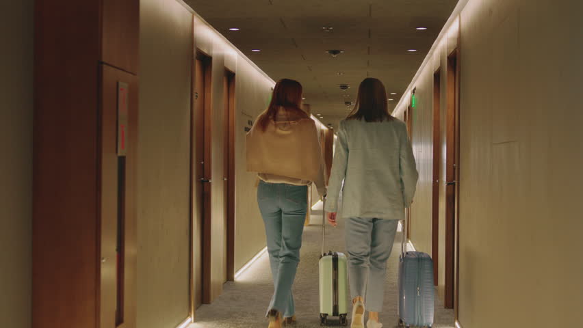 Back view of two slim ginger and blonde businesswomen in elegant outfits walking in modern hotel corridor. Women rolling suitcases. Gorgeous interior. Distance shot. Atmosphere of elegance and luxury Royalty-Free Stock Footage #1108011571