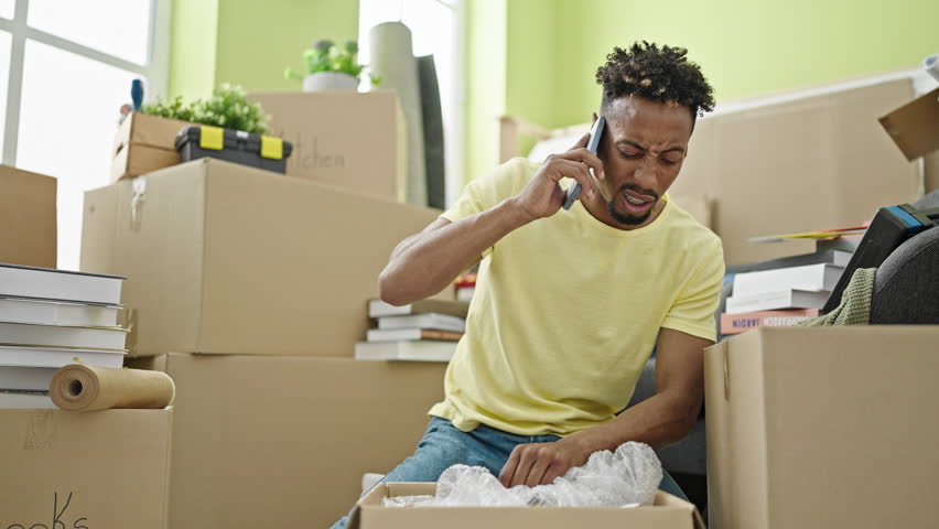 African american man unpacking cardboard box talking on smartphone looking upset at new home Royalty-Free Stock Footage #1108018517