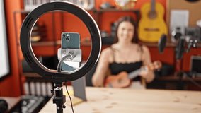 Young beautiful hispanic woman musician recording video tutorial playing ukulele at music studio