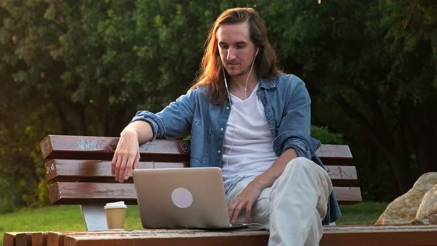 Young man watching video on his laptop and drinking coffee while sitting at public park at sunset | Shutterstock HD Video #1108025223