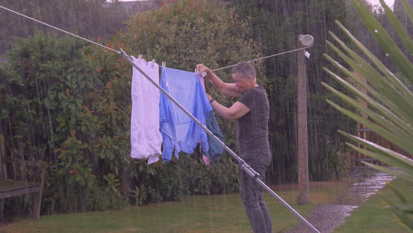 A senior woman rushing to unpeg clothes on a washing line in pouring rain in a garden or yard, then throwing the pegs and clothing into baskets, before picking them up and hurrying away. Royalty-Free Stock Footage #1108041341