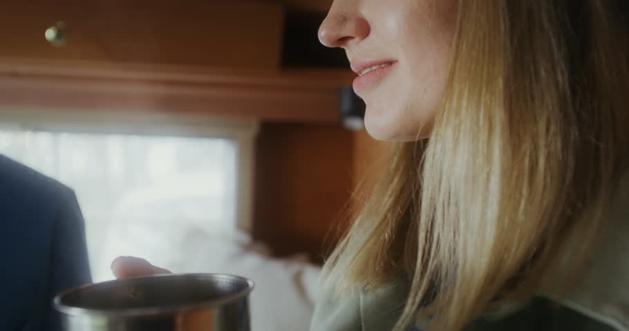 A young woman in a hat drinks a hot drink from a metal mug while sitting in a RV. She smiles and looks at the person sitting next to her