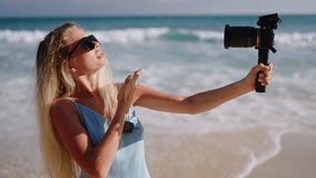 Woman travel vlogger adores summer vacation in Sri Lanka at ocean beach filming herself on camera. Young blonde blogger in sunglasses walking on sand holding professional movie camera. Slow motion.