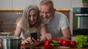 Laughing retired old gray haired Caucasian family love couple using mobile phone online order delivery food vegetables on kitchen table happy senior woman and man talking shopping cooking healthy meal