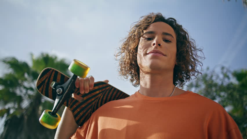 Happy trendy skateboarder smiling camera holding modern skateboard tropical palms. Portrait curly teenager skater posing sunny nature with longboard. Cheerful relaxed guy model in sunlight close up