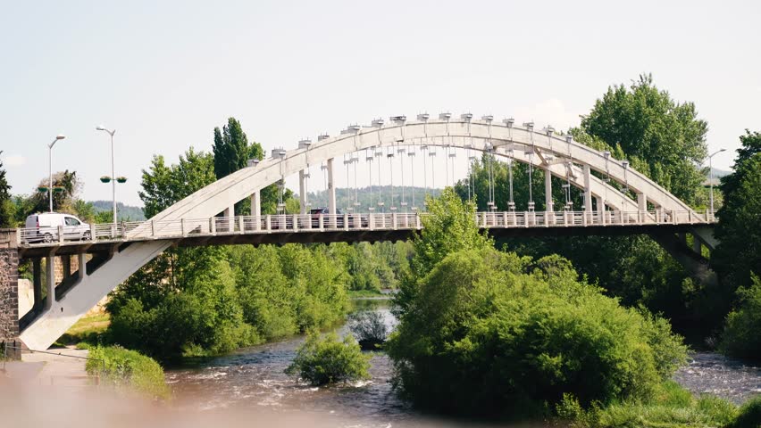 Side view of a white arched bridge across a mountain river along which cars drive. The commune of Langeac in the Haute-Loire department in France