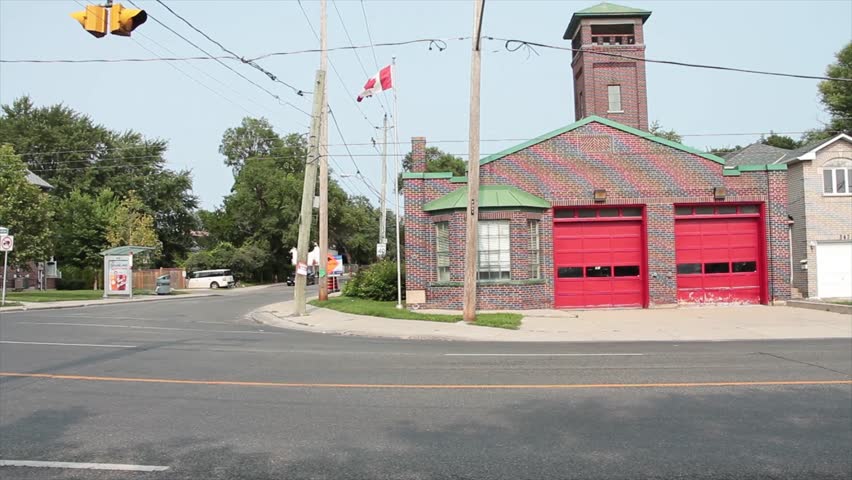 fire department station small boutique vintage brown burgundy brick with two red garage doors and steeple and canadian flag blowing waving on flagpole with road with light traffic, wide shot