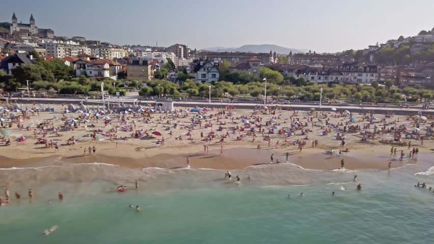 Aerial view tracking left above Ondarreta Beach in La Concha Bay, San Sebastian, Spain. White sandy beach crowded with sunbathers as seagulls fly past.
