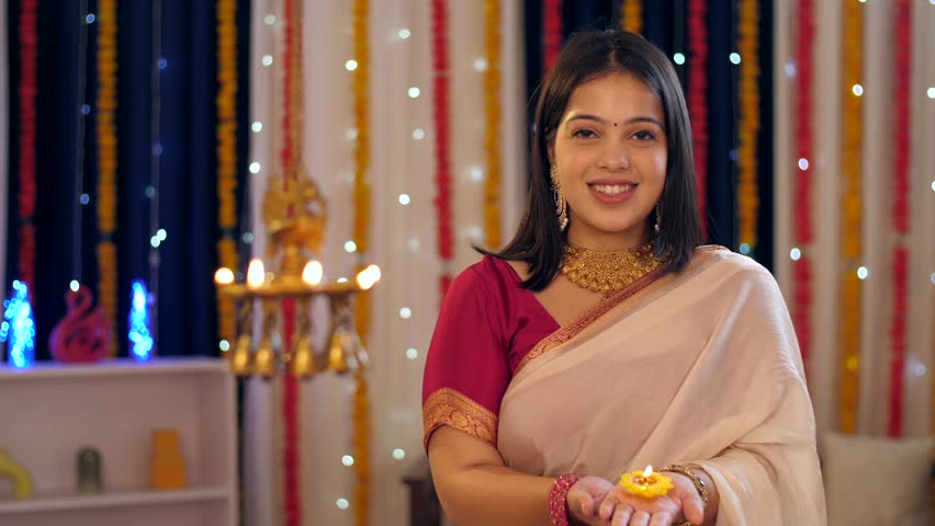A beautiful woman holding a brightly burning clay diya to celebrate Diwali - greetings, posing for camera. A pretty lady in ethnic clothes posing for the camera against a festive background - India...