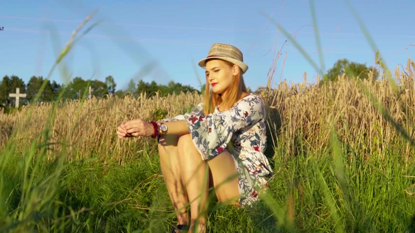 Stunning HD footage of a white caucasian girl in a dress with a knitted hat and red lipstick sitting in a wheat field, lost in thought, basking in the setting sun