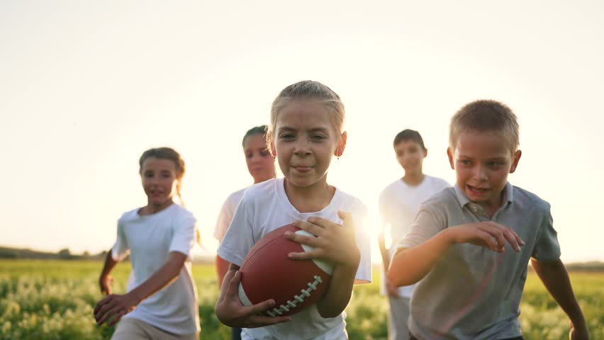Happy team of children in park at sunset. Children play rugby ball in american football in park. Summer games children in park in nature run on the green grass. Family plays runs in team games in park