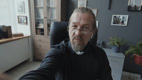 Handheld POV of modern mature Catholic pastor sitting on chair in his office talking to someone on video call