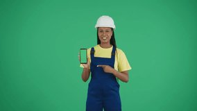 Medium green screen isolated chroma key video of a dark-skinned young female construction worker holding a phone with advertising area, workspace mock up, pointing at it and speaking.