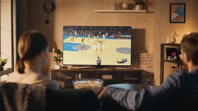 Young Couple Spending Time at Home, Sitting on a Couch and Watching and Exciting Live Basketball Tournament on TV in Their Stylish Loft Apartment. Man and Woman Eating Snacks and Enjoy the Sports Show - Powered by Shutterstock - Get 15% off with code: PIKWIZARD15