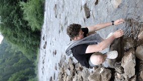 a guy in a black t-shirt and shorts washes his face in a mountain river. a man wipes his face with a towel against the background of mountains and forest. vertical video