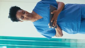 Vertical video portrait of smiling mature female doctor wearing scrubs checking digital tablet in hospital - shot in slow motion