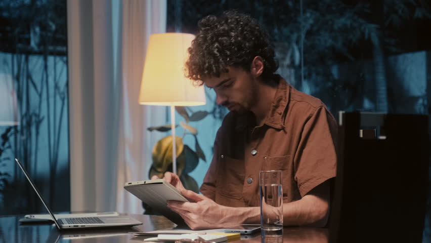 Young male entrepreneur working on digital tablet and laptop sitting at table in living room at night time