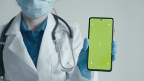 A medical specialist uses a smartphone to present medical services and advertise pharmaceuticals. The doctor points his hand to the phone screen. The effect of chromakey on the green screen gadget