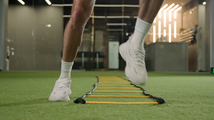 Unknown professional footballer running exercise jumping between ladder sticks male sportsman man soccer player muscular legs in sneakers fast jogging in sport gym training workout on football pitch