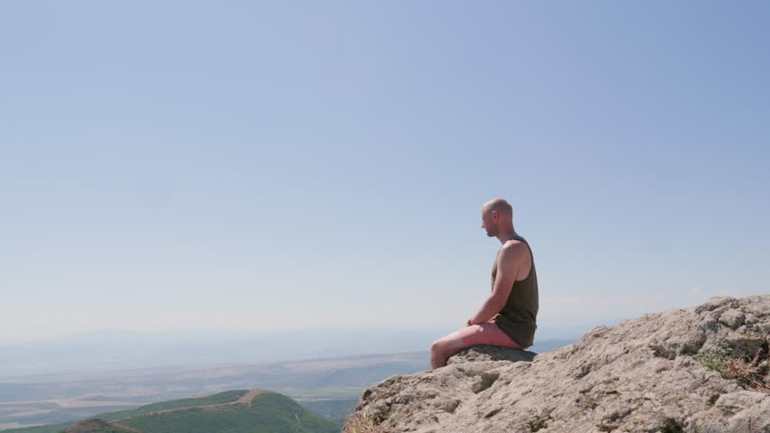 one guy sits on the edge of a cliff and dangles his hanging legs, he looks around and jumps down, dressed in a T-shirt, shorts and sneakers, on his hand is a gold watch