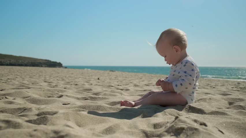 Active, cheerful toddler plays along a sandy sea beach on sunny summer day. Slow motion: Baby is playing on the sea beach on sunny day. Little boy exploring the world on sand beach against  a sea Royalty-Free Stock Footage #1108115263