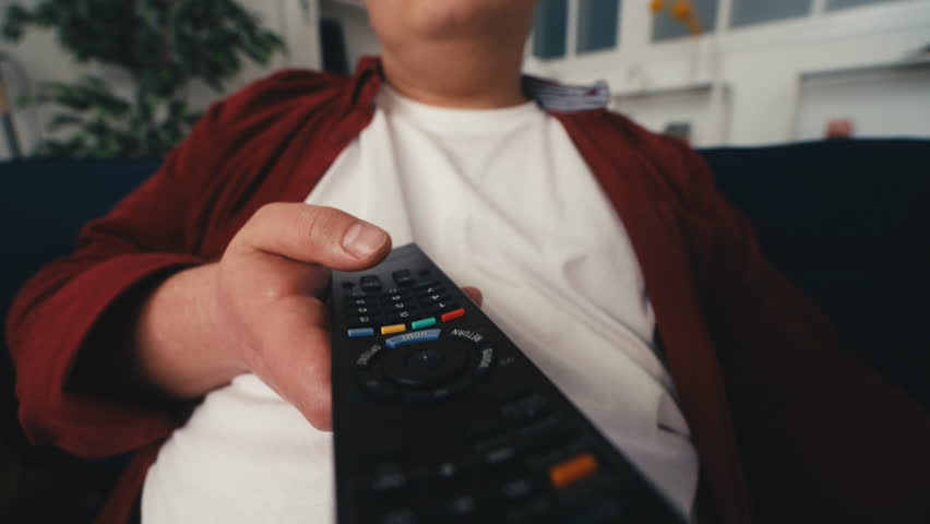 Young overweight man watching TV and drinking beer on couch, unhealthy lifestyle