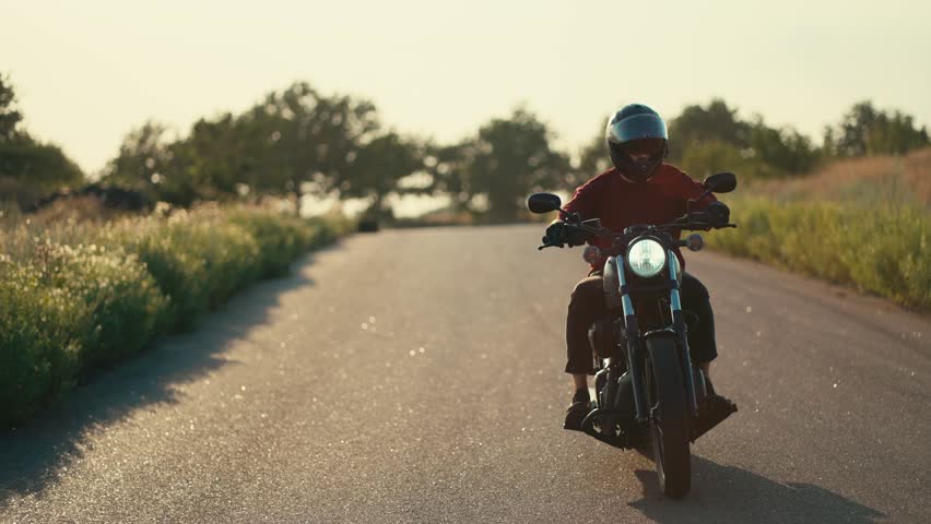 A motorcyclist in a red jacket and in a helmet is riding on a road in sunny weather with a breeze