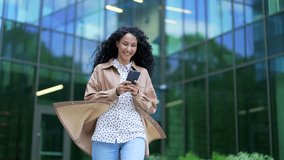 Young smiling businesswoman is using a smartphone walking on the street near an office building. Happy female employee chatting online, texting with a colleague or boyfriend, browsing social media - Powered by Shutterstock - Get 15% off with code: PIKWIZARD15