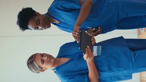 Vertical video of two smiling mature female doctors wearing scrubs meeting and checking digital tablet in hospital - shot in slow motion