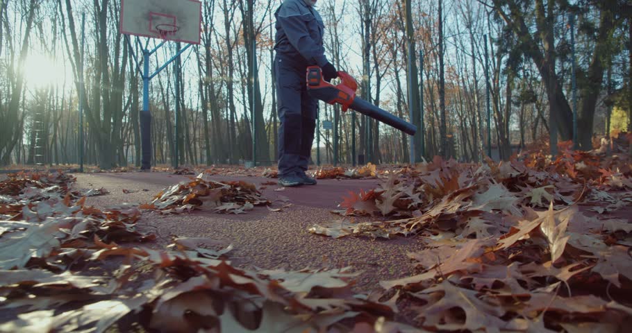 Grey haired caucasian man cleaning autumn park with leaf blower. Senior concierge in uniform, gloves and headphones removing fall foliage at city area. Royalty-Free Stock Footage #1108121417