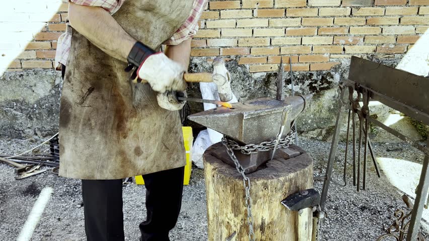 Blacksmith turns and hammers red hot metal piece against anvil chained to wooden log