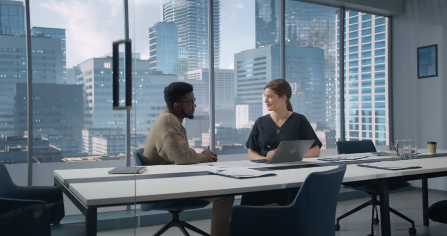 Two Multiethnic Colleagues Have a Discussion in Bright Modern Skyscraper Office in an Agency. Balck Creative Director and Female Project Manager Talking About Data On Laptop Computer.