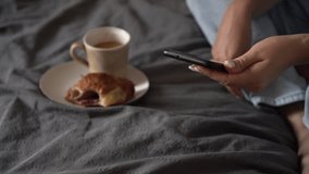 Woman is looking for information in a smartphone  and drinking coffee with a croissant. Watch videos, movies, series on your phone without leaving your home. Cozy house, free time, coffee time 