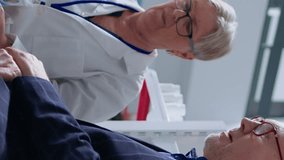 Vertical video Elderly pharmacist in drugstore using electronic thermometer on senior man, checking temperature. Old patient and healthcare specialist during yearly examination, monitoring flu