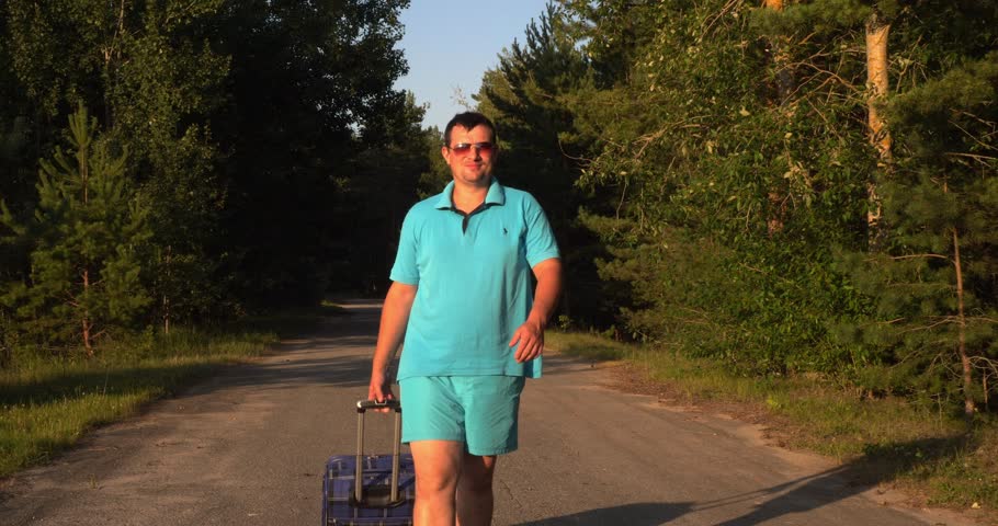 A male tourist walks with a suitcase at sunset along the road to the hotel located in the forest. Camping . A man in sunglasses came to relax in nature in an eco hotel.