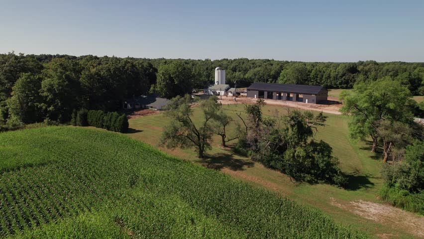 Cornfield and farm land in field next to large barn, storage shed, and tall silos from a drone establishing aerial shot above farming building structure in Midwestern rural area of New Albany, Ohio