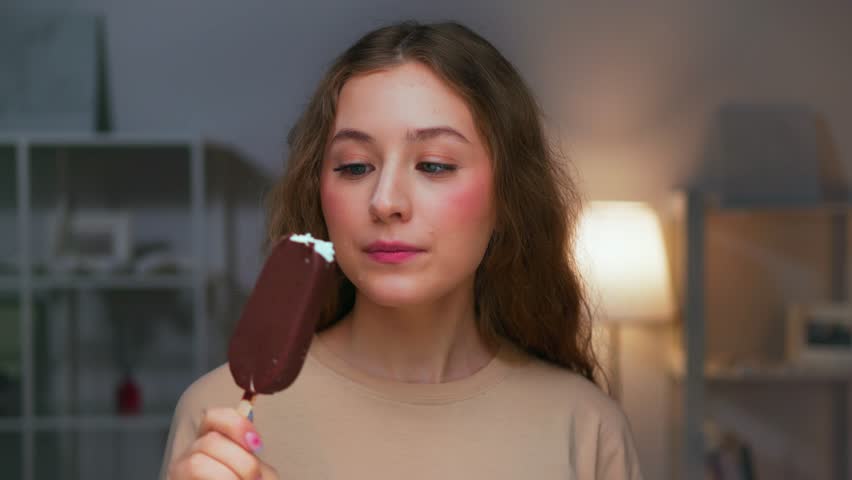 Portrait of Young Woman Eating Sweet Food as Looking at Camera. Real Person on Diet Consuming Milk Ice Cream as Lunch Snack. Attractive Female Human Inside Cozy Living Room Alone with Sugary Milky Bar Royalty-Free Stock Footage #1108147325