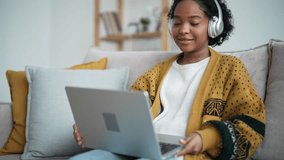 Portrait african american woman in earphones has video call on laptop sits on couch at home. Friendly smiling girl talking waving hand looking at screen. Online conversation, communication concept.