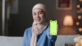 Smiling covered Muslim woman in hijab showing smartphone screen with chroma key. Advertisement. Video for insert promotional clip movie. Cell phone device with green motion tracking points. 