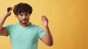 Video portrait of a handsome curly-haired young stylish Indian man in a turquoise t-shirt fixing his curly hair and looking at the camera as a mirror while standing in a studio on a yellow background.