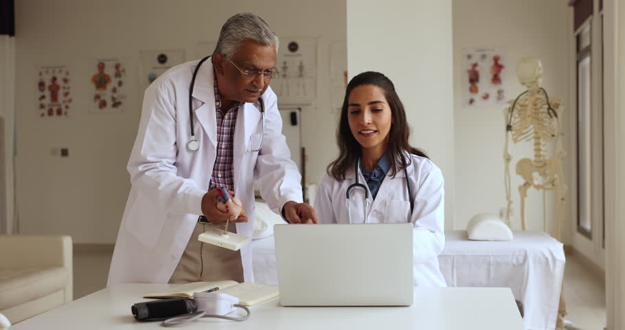 Indian and Latina cardiologist colleagues work together in clinic office, using laptop discuss telemedicine platform, old experienced GP teach intern at meeting in hospital. Internship, apprenticeship Royalty-Free Stock Footage #1108158111